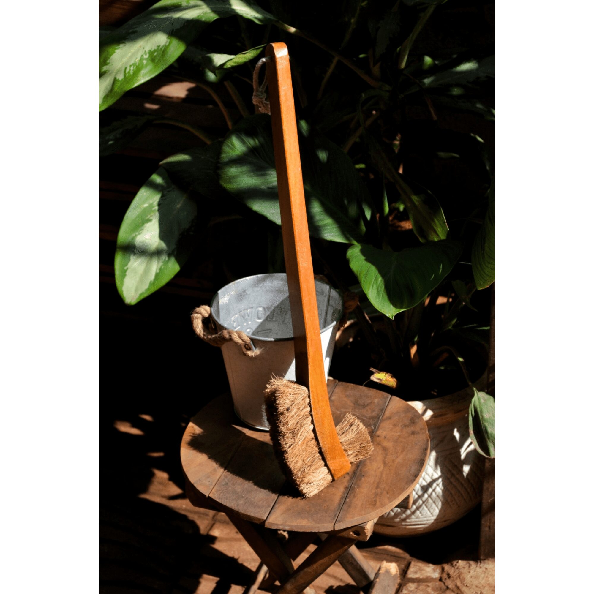 Natural coconut coir double hockey toilet brush with long wooden handle, placed on a rustic table beside a metal bucket.