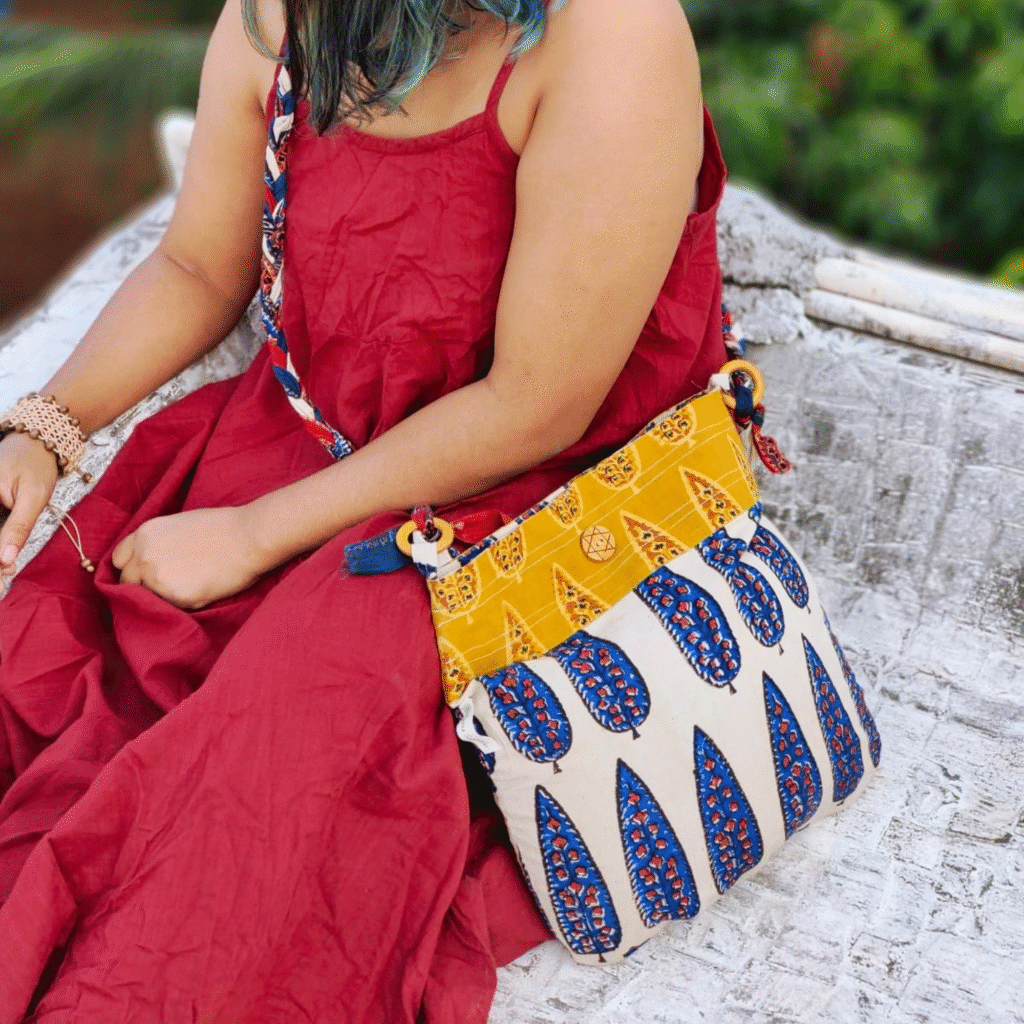 A stylish woman in a red dress holding a sustainable, hand block printed cotton sling bag featuring a braided multicolor strap and traditional blue and mustard ethnic patterns, crafted by Almitra Sustainables. The bag highlights eco-friendly, artisanal craftsmanship using natural dyes and ethically sourced materials.