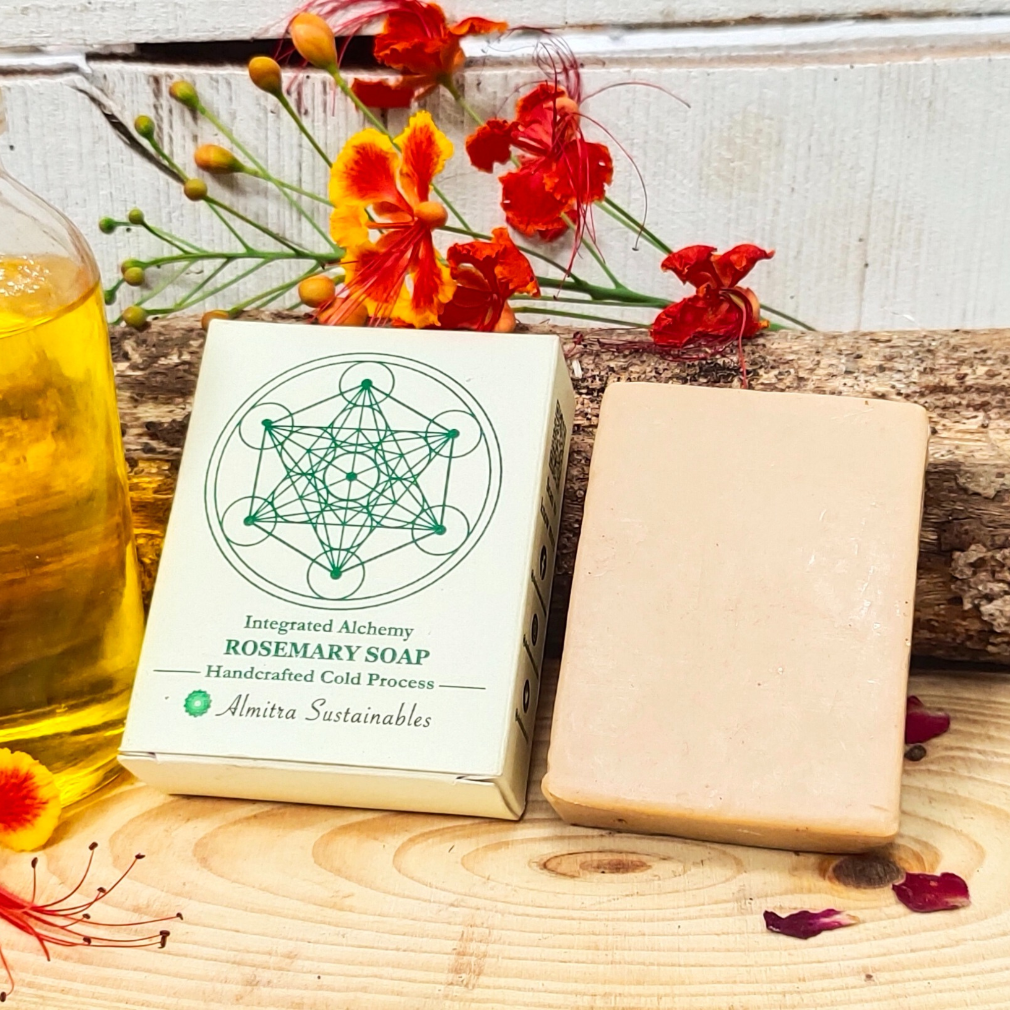 A handcrafted rosemary soap bar by Almitra Sustainables, made using the traditional cold process method. The soap is shown resting on a raw wooden slab, alongside its eco-friendly paper packaging printed with green sacred geometry. Surrounded by vibrant red-orange flowers and a glass bottle of herbal oil, the display evokes a natural, sustainable, and plastic-free skincare experience, ideal for conscious consumers seeking zero-waste bath products.